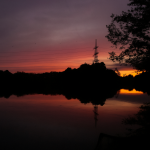 Sonnenuntergang über einem ruhigen See mit Spiegelung im Wasser, Silhouetten von Bäumen und einem Strommast im Hintergrund.